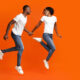 couple young man and woman holding hands, looking at each other and running in the air towards copy space over orange studio background