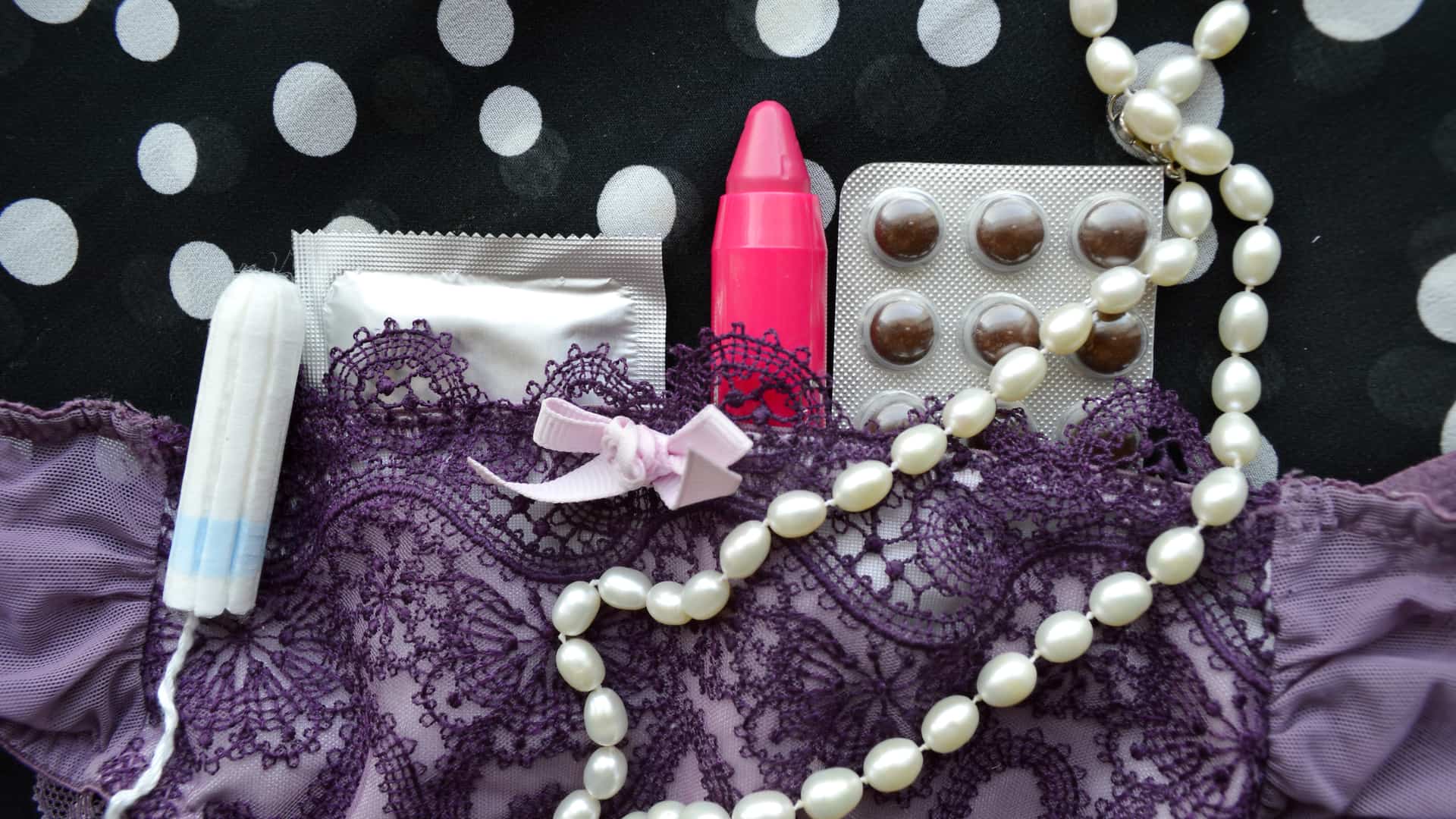 Closeup oral contraceptive pills, tampon, condoms and pink lipstick on lace lingerie.