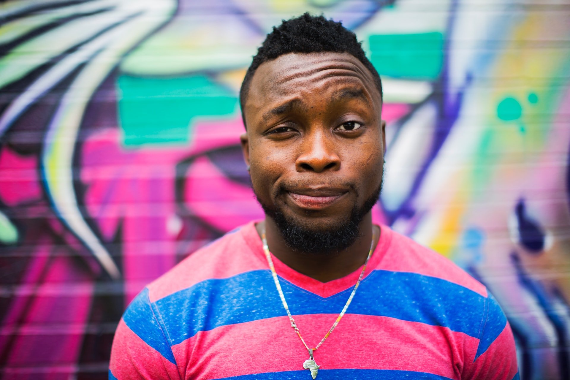 A black man in a colorful t-shirt and chain, raising his eyebrow at the camera