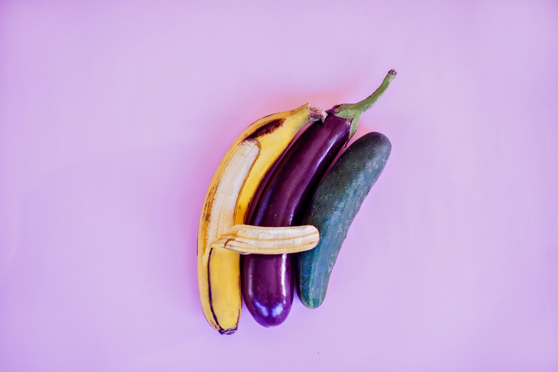 A banana, eggplant and cucumber next to each other, with one banana peel reaching across the other two fruits