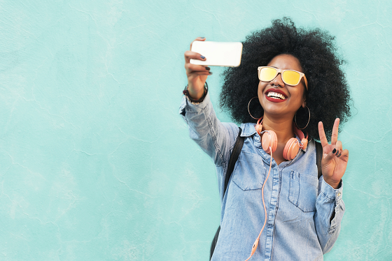 Women with an afro, yellow glasses, and headphones around her neck taking a selfie