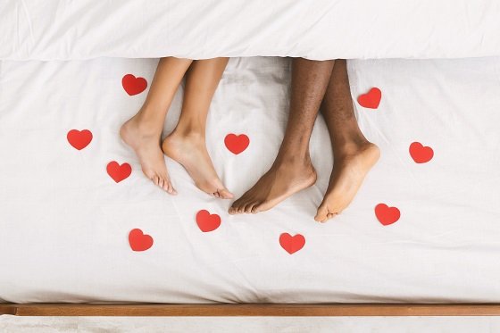 two pairs of feet on edge of bed surrounded by red love symbols