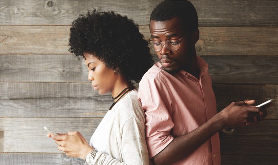 Couple standing back-to-back, with him peeking at her phone 