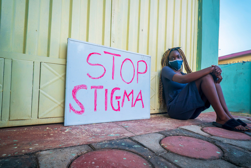A beautiful young black woman, wearing a locally made mask sitting beside a placard educating on stop stigma