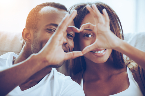 Beautiful young African couple sitting close to each other and looking through a heart shape made with their fingers